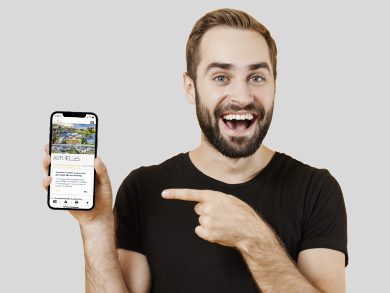 Mann mit Bart, der ein Smartphone in der Hand hält. Auf dem Handy ist die Regio-App der Gemeinde Linkenheim-Hochstetten