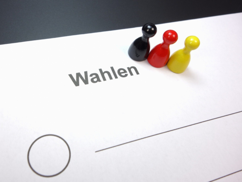 Stimmzettel für eine Wahl mit 3 Spielfiguren aus Holz in den Farben schwarz, rot und gelb